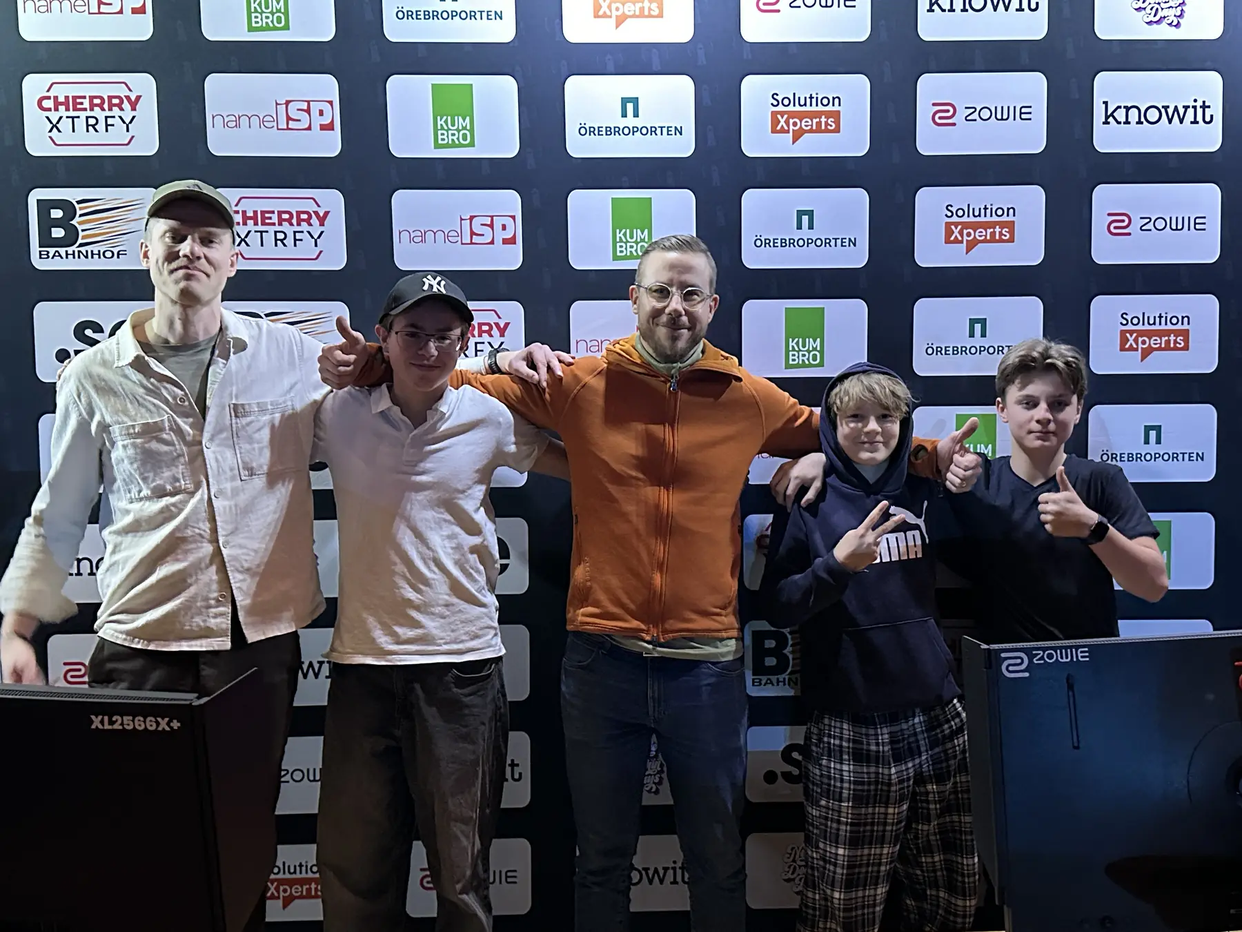 Fem deltagare står med armarna om varandra framför sponsorväggen efter en turneringsfinal.